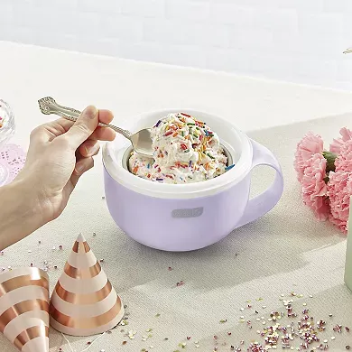 A white Dash Mug Ice Cream Maker filled with ice cream, next to pink party decorations and a silver spoon inside the mug for scooping.