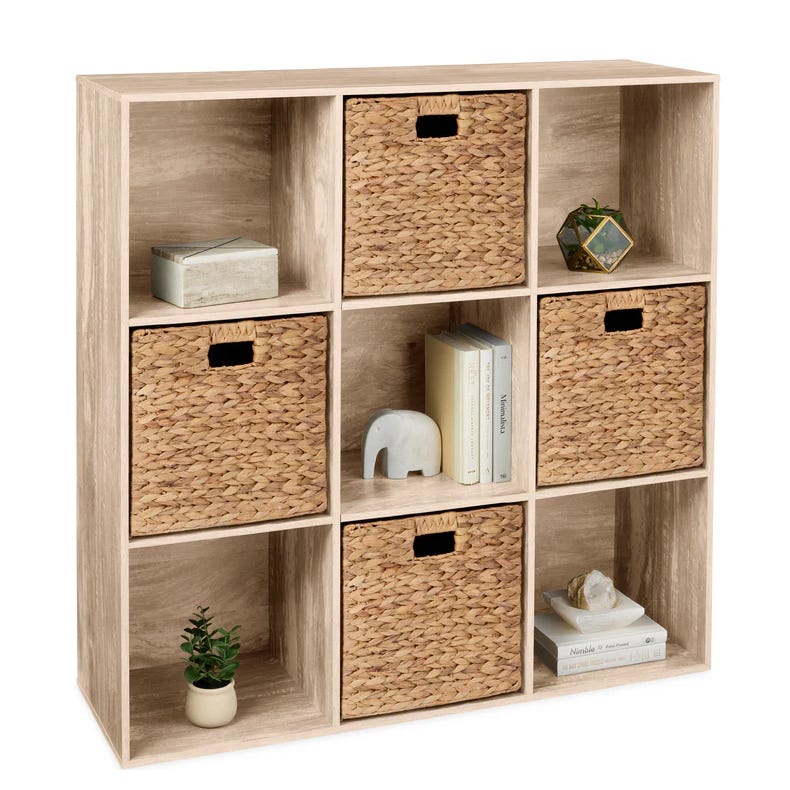 A 9-cube bookshelf with a light wood finish featuring four woven baskets, three books, a small potted plant, a geometric terrarium, a decorative box, and an elephant figurine. Three cubes are empty, and dimensions are not specified.