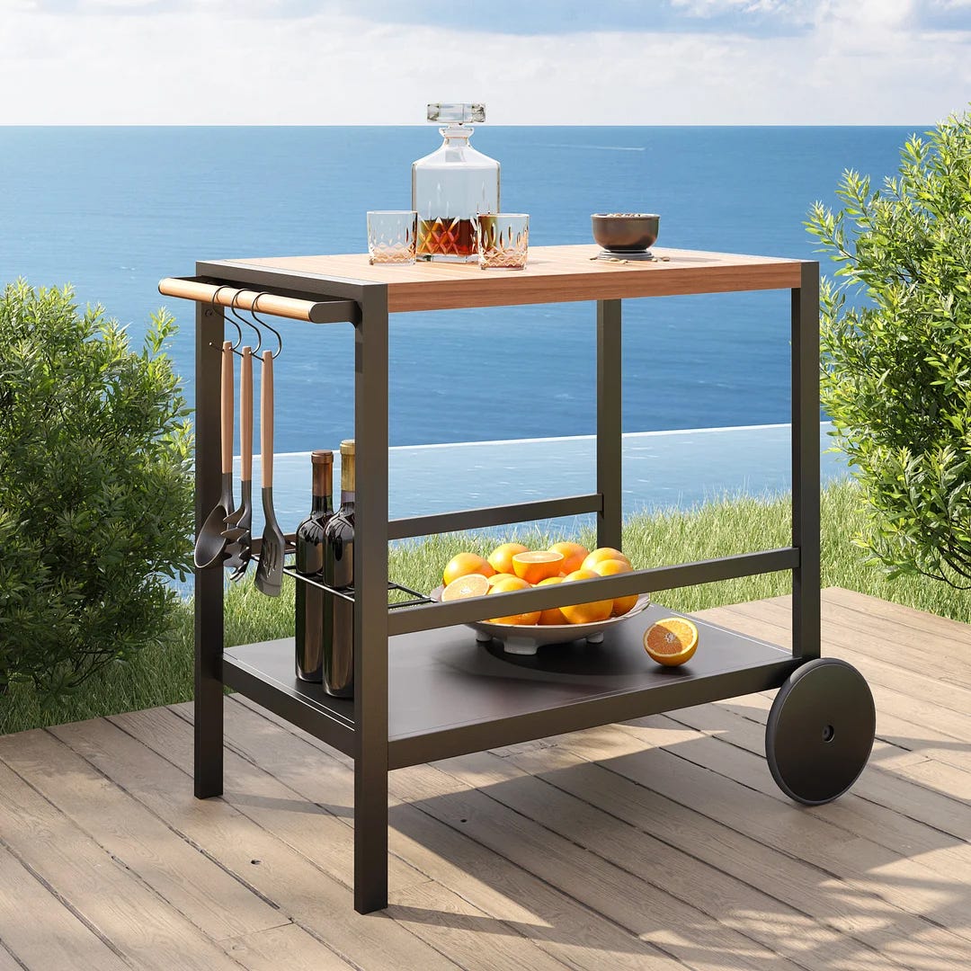 A portable outdoor kitchen cart with a wooden top, storage shelf, hanging utensils, and wheels. There's a decanter, bowl, and fruit on it.