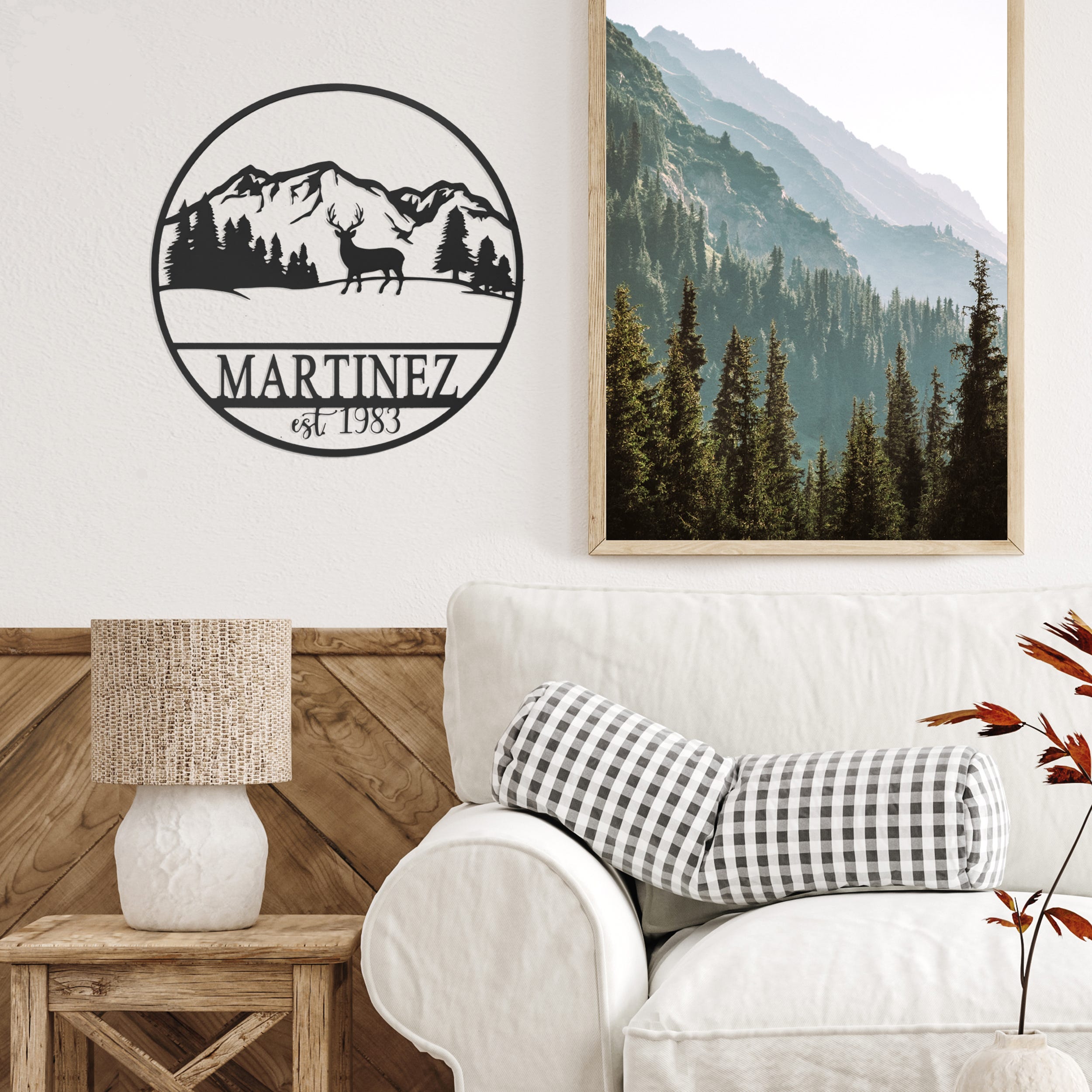 A personalized wall sign beside a framed scenic forest photograph, above a side table with a lamp, and a white armchair with a checkered pillow.