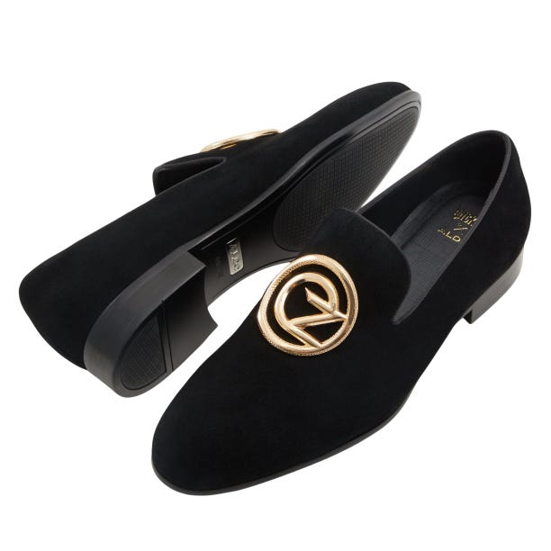 Black suede loafers with a low heel, featuring a gold, circular emblem on the front. The inner sole is branded, and the outer sole has a textured grip.