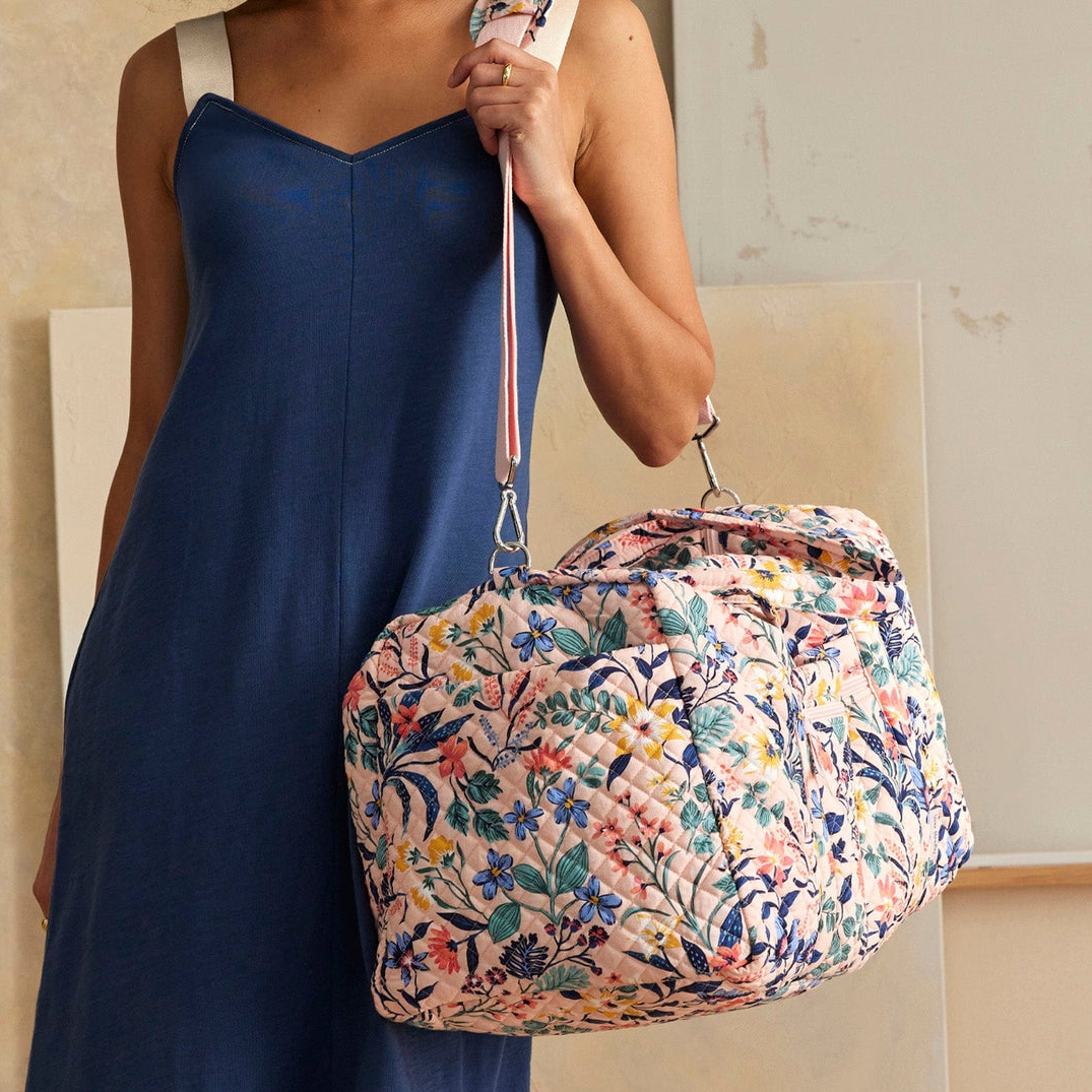 A floral quilted duffel bag with pastel colors and an adjustable strap held by a person in a blue dress.