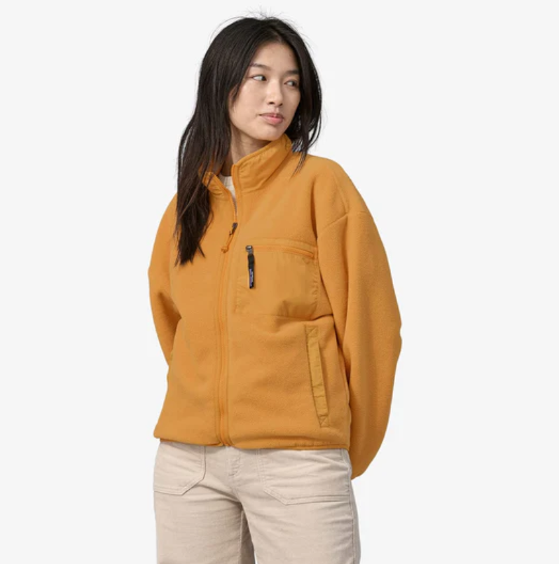 A woman is wearing a mustard yellow fleece jacket with a zippered chest pocket and light beige pants.