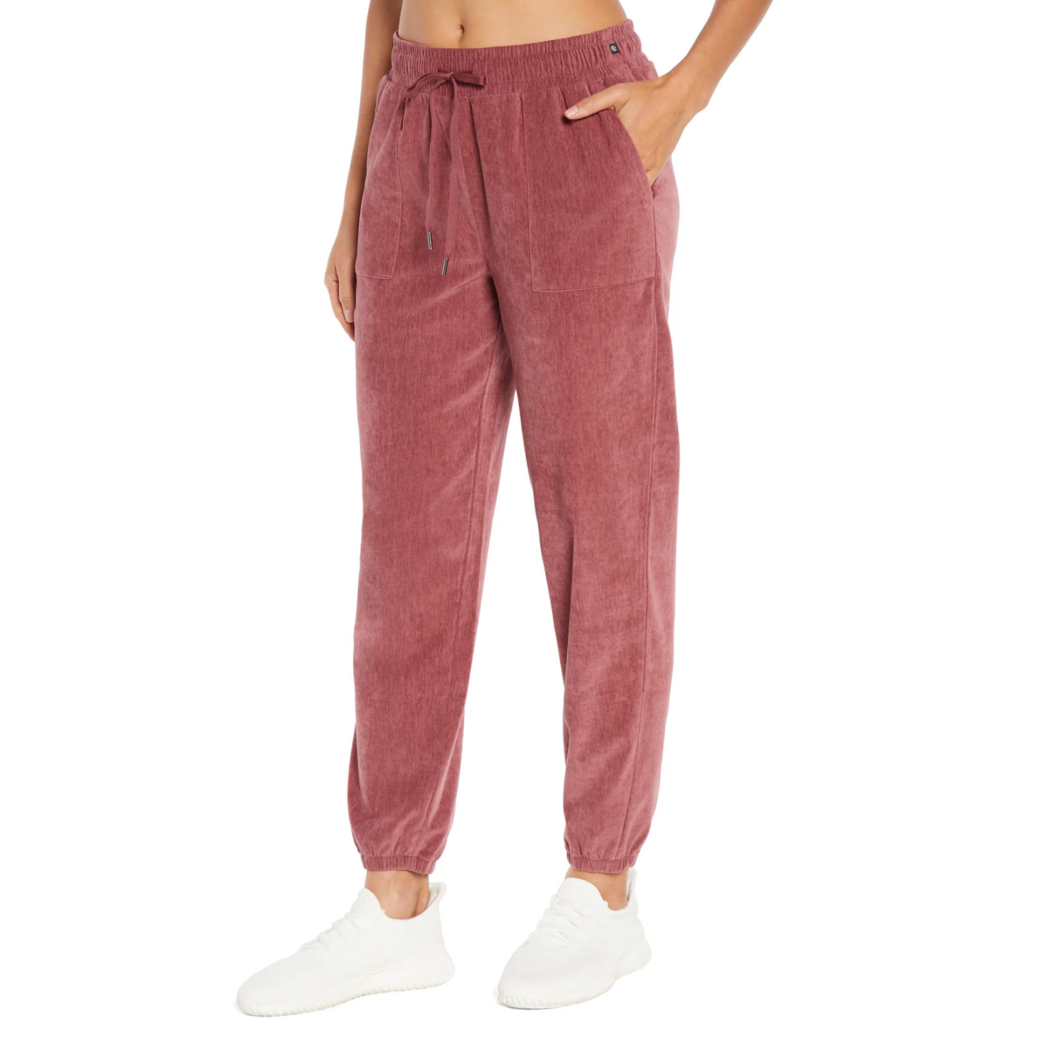The Marika Solana Joggers are pink, made of soft, textured fabric. They feature an elastic waistband with a drawstring, side pockets, and elastic cuffs, paired with white sneakers.