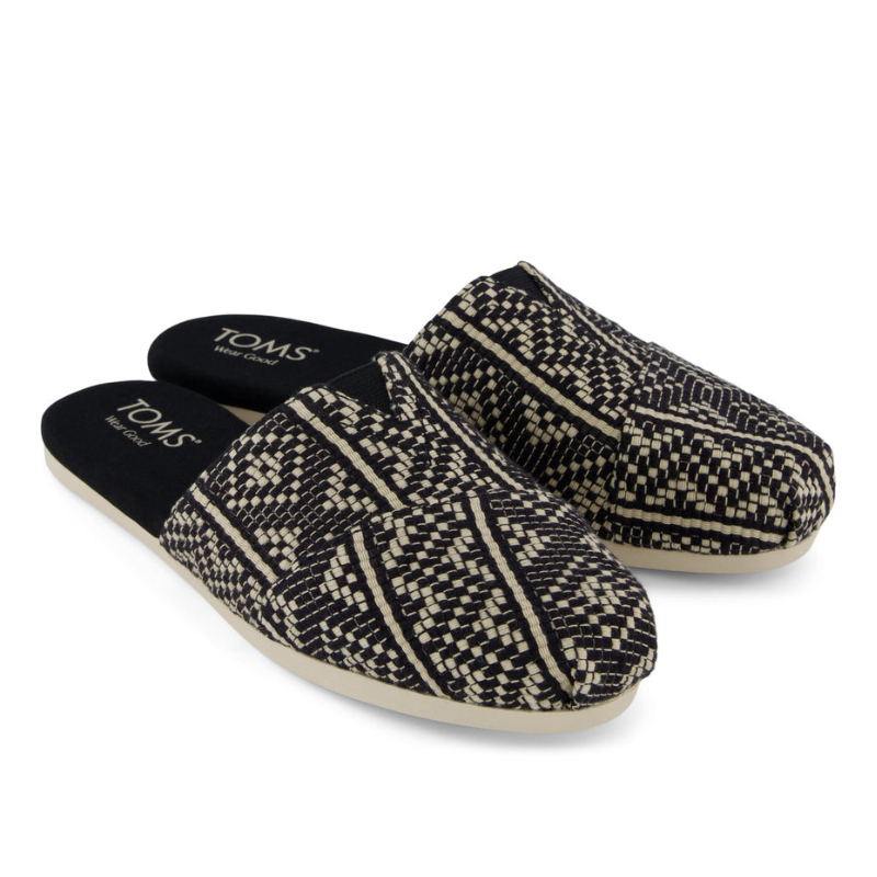 A pair of black and white patterned slip-on shoes with a closed toe and open heel design, featuring the brand \“TOMS\“ on the insole.