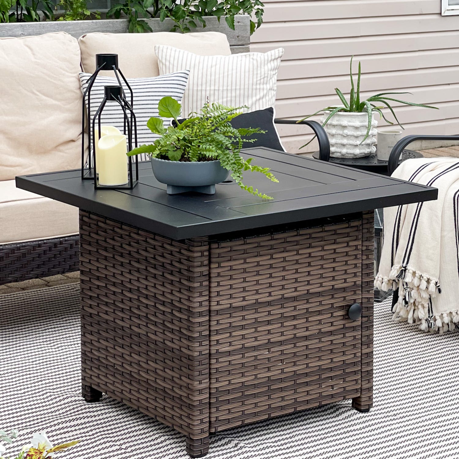 A wicker-style patio fire pit table with a black top is shown, featuring a potted plant and a lantern with a candle. It is set on a striped outdoor rug, next to a sofa with cushions and a chair with a potted plant.