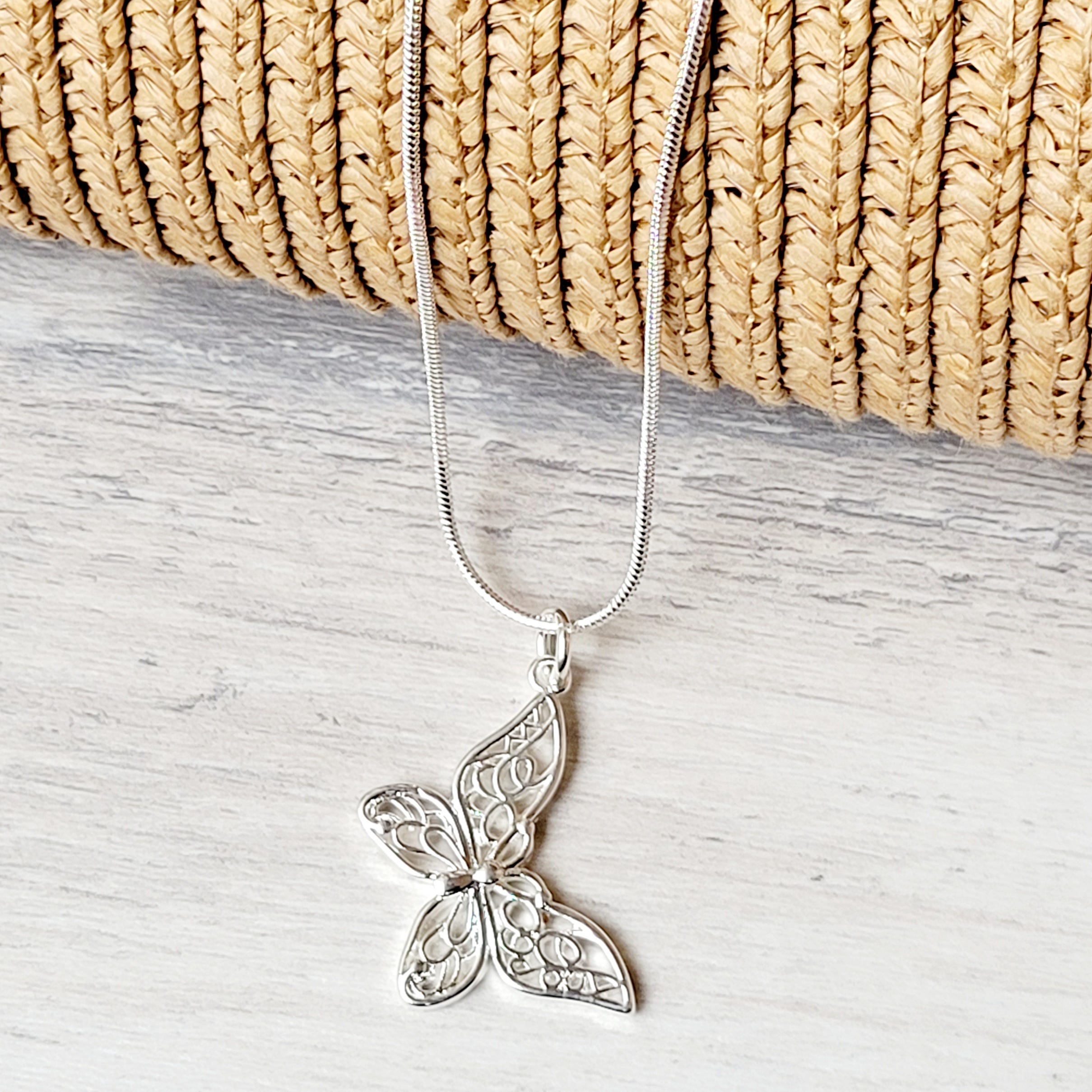 Silver butterfly pendant on a necklace chain, displayed against a woven basket and wooden surface.