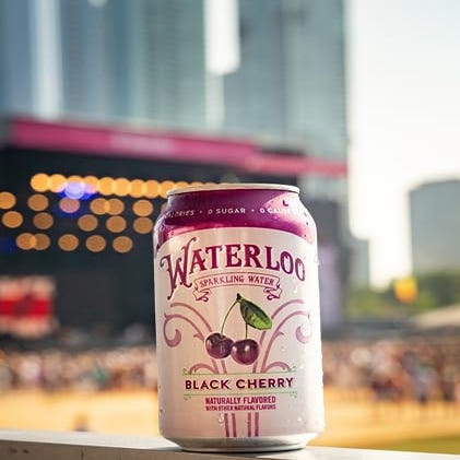 A can of Waterloo Sparkling Water with Black Cherry flavor is displayed with a blurred background of a crowded event and city buildings.