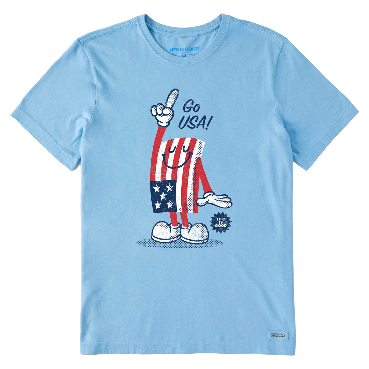Light blue T-shirt featuring a cartoon flag character with a smiley face, wearing white shoes, pointing upward with a finger, and the text \“Go USA!\“ above. The design incorporates red, white, and blue colors with stars, and the \“Life is Good\“ logo.