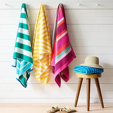 Beach towels with colorful stripes and dots hanging on hooks beside a wooden stool with a straw hat and sandals below.