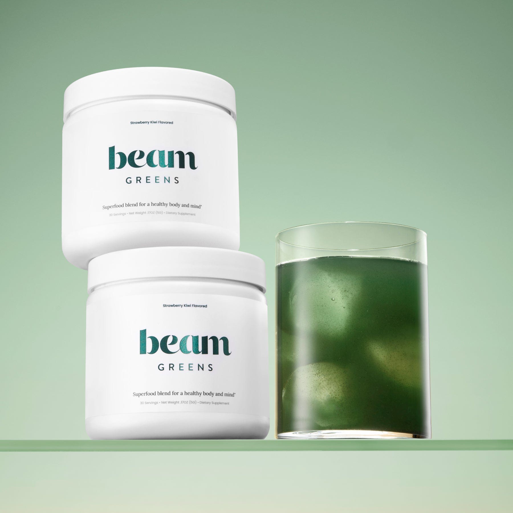 Two white containers labeled \“beam GREENS\“ are beside a glass filled with a green liquid, suggesting a greens supplement drink mix.
