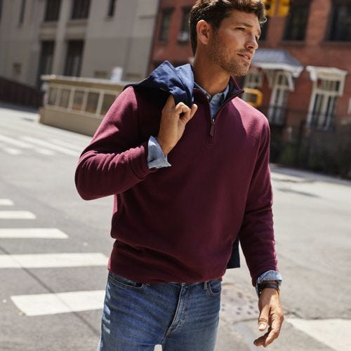 A man wears a burgundy quarter-zip sweater with a raised collar over a blue shirt paired with casual denim jeans, carrying a navy jacket over his shoulder.