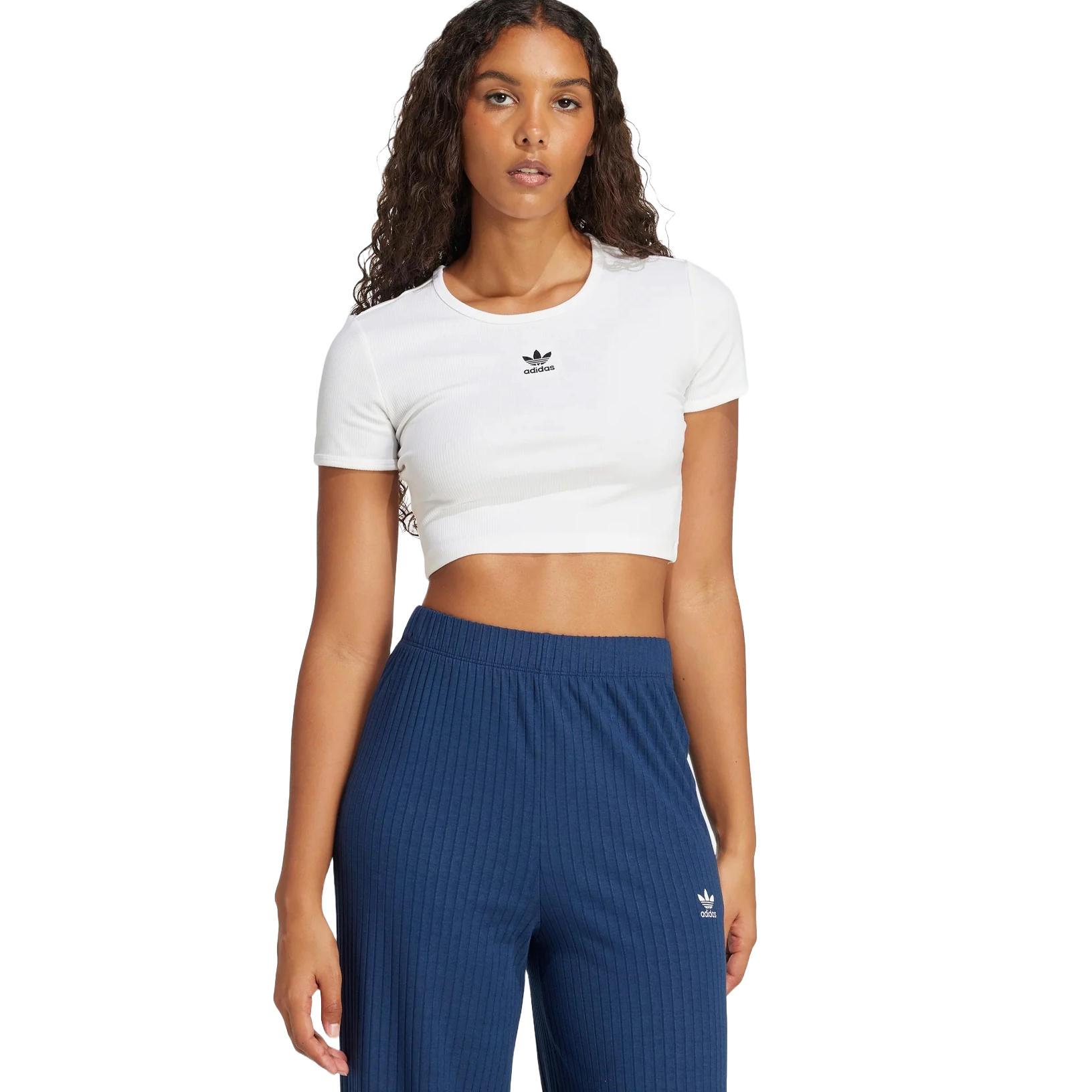 A woman wears a white Adidas cropped t-shirt with a logo on the chest and blue ribbed Adidas pants, both featuring the trefoil logo.
