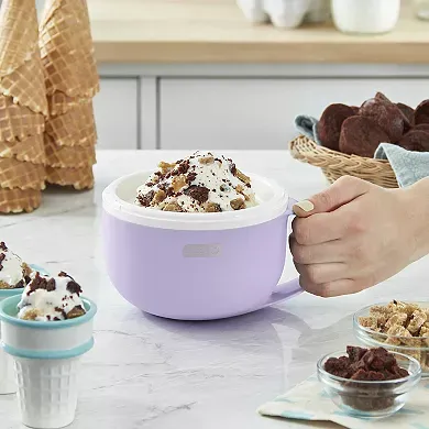 A purple Dash mug ice cream maker is on a kitchen counter with a hand scooping out freshly made ice cream topped with sprinkles and cookies.