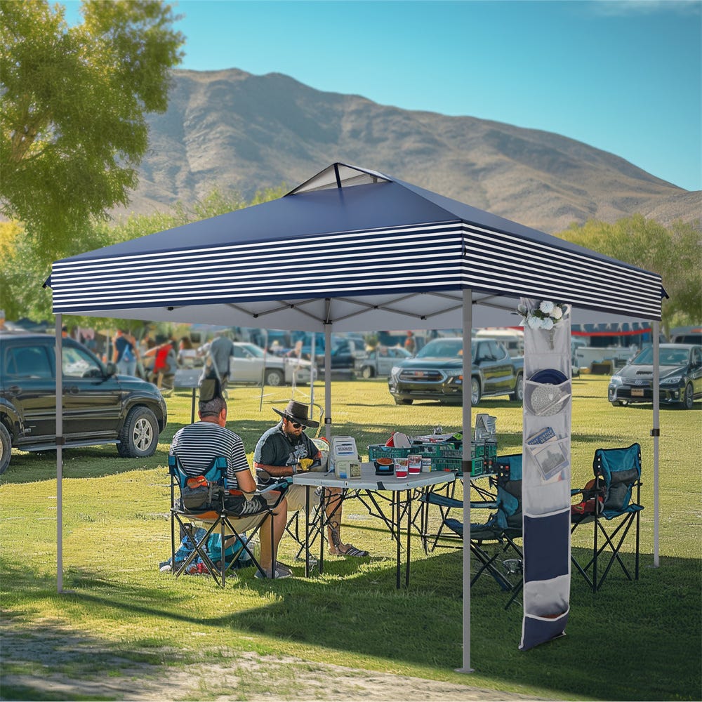 A striped canopy tent set up in a grassy area with two people sitting at a table underneath, surrounded by outdoor gear and vehicles in the background.