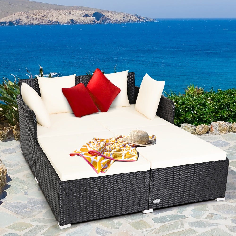 Outdoor patio furniture set with white cushions, red accent pillows, and a black wicker frame, by the sea.