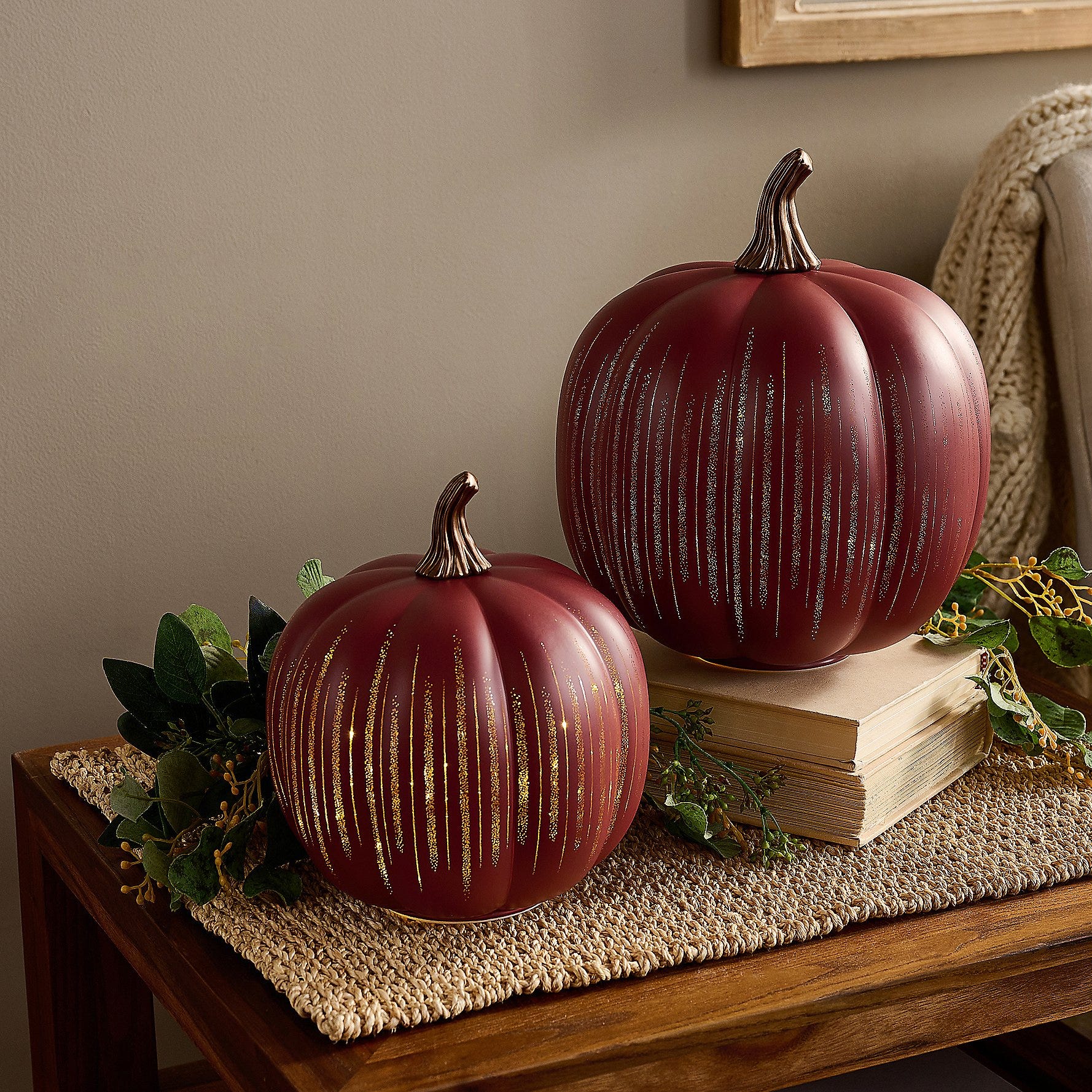 Two decorative red pumpkins with vertical metallic stripes, one larger and one smaller, sit on a woven mat. They are accompanied by faux greenery and books for an autumn-themed display.