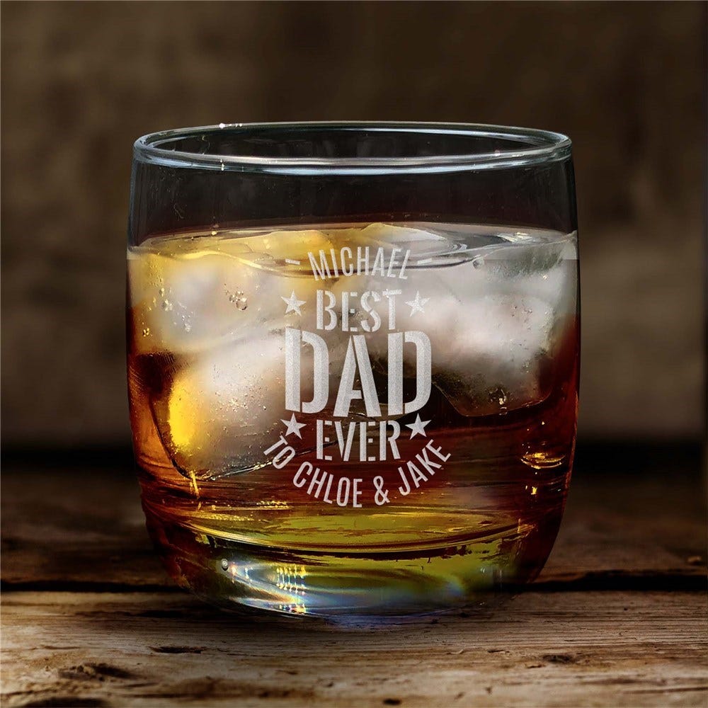 A whiskey rocks glass with the engraving \“Michael, Best Dad Ever, to Chloe & Jake,\“ filled with an amber liquid and ice cubes, set on a wooden surface.