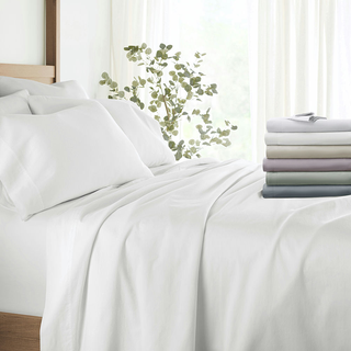 White bedding set displayed on a bed with a stack of folded sheets in various colors at the foot, next to a potted plant.