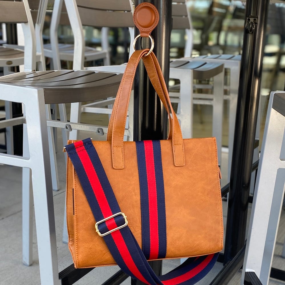 A brown leather tote bag with a magnetic bag hanger attaches to a metal bar. It features a colorful, adjustable blue and red striped shoulder strap with a gold buckle for secure carrying on tables or bars.