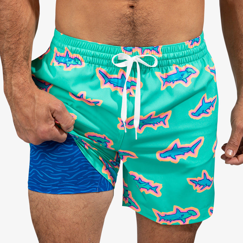 The swim trunk features a vibrant shark print on a turquoise background with a white drawstring. It includes a blue inner lining with a wave pattern for added comfort and support.