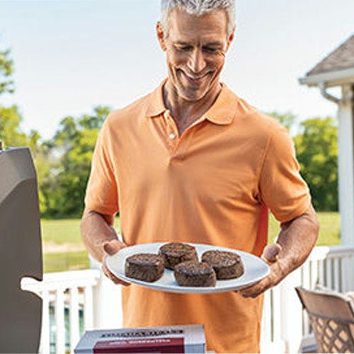 A man holding a plate with six grilled burgers next to an outdoor grill.
