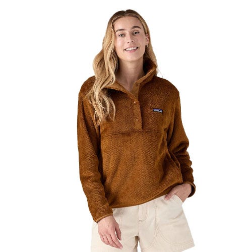 A woman models a brown Patagonia half-snap pullover with a cozy fleece texture and a front chest logo. She pairs it with white shorts, her hand in one pocket, conveying a casual, comfortable outdoor look.