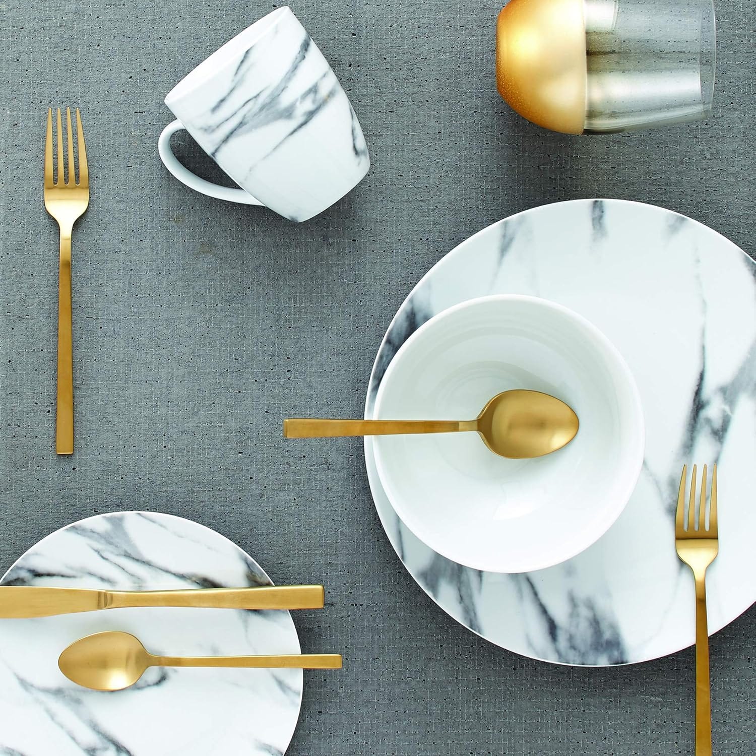 Marble-patterned dinnerware set with a mug, plates, and bowl paired with gold-toned cutlery on a textured gray surface.