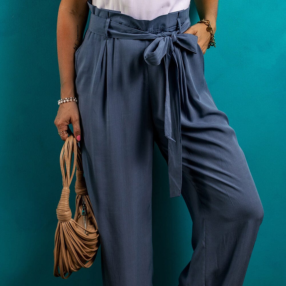 Person holding a tan knotted handbag while wearing blue high-waisted trousers with a bow tie at the waist.