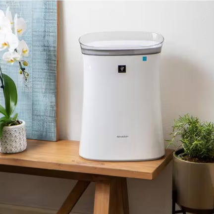 White cylindrical air purifier on a wooden table, flanked by potted plants.