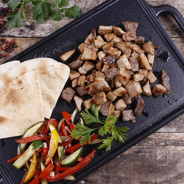Sliced grilled meat, pita bread, and sautéed vegetables on a cast iron pan, seasoned with herbs and spices.
