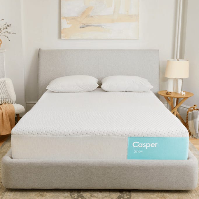 Casper Snow Mattress is a bed with a light gray upholstered frame and a white mattress featuring a smooth, dotted texture. It includes two white pillows and is situated in a minimalistic room with a side table and lamp.
