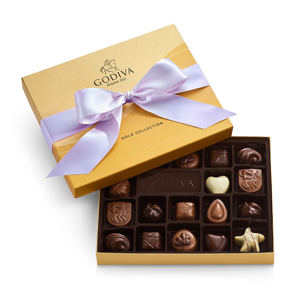 A Godiva Gold Collection box featuring 16 assorted chocolates, elegantly wrapped in a gold box with a lavender ribbon. The assortment includes various shapes and flavors in milk, dark, and white chocolate.