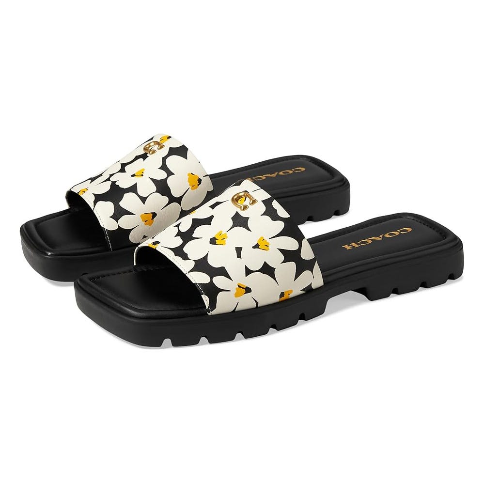 A pair of black and white floral print slides with a scalloped pattern and a gold-tone emblem on the upper strap.