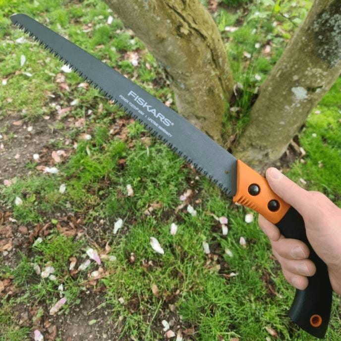 A hand holding a Fiskars saw with a black and orange handle, used for cutting wood.