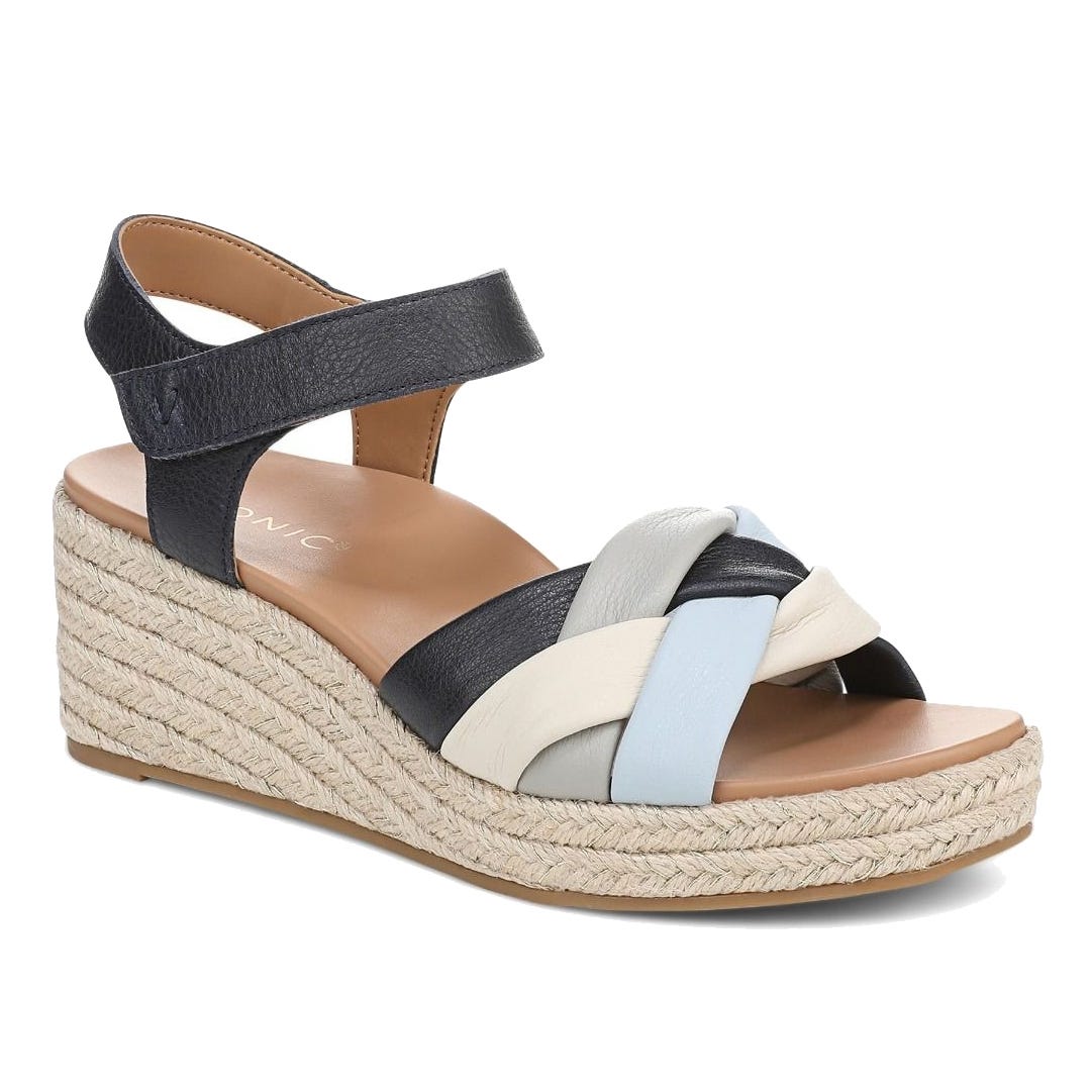 A wedge sandal with a braided multi-color strap design, featuring a mix of black, white, and light blue. It has a jute-wrapped platform sole and an adjustable ankle strap.