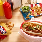 A variety of Fritos corn chips are scattered around a hot dog topped with chili and jalapeños, flanked by condiment bottles and a bowl of sliced jalapeños on a wooden table.