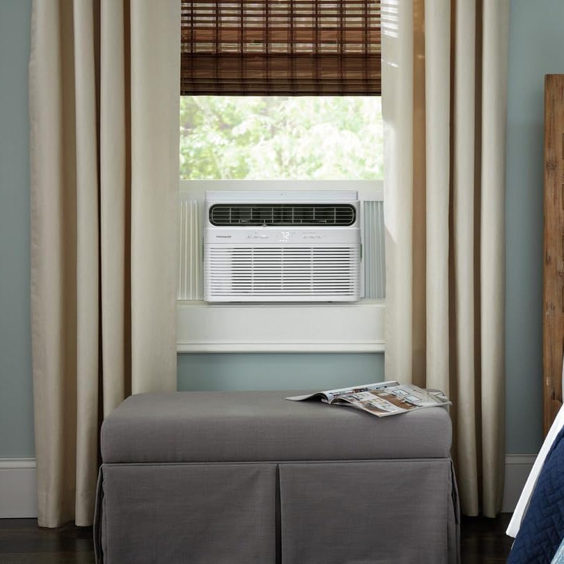 Window-mounted air conditioner installed above a bench with a magazine on it, beside beige curtains and bamboo shades.