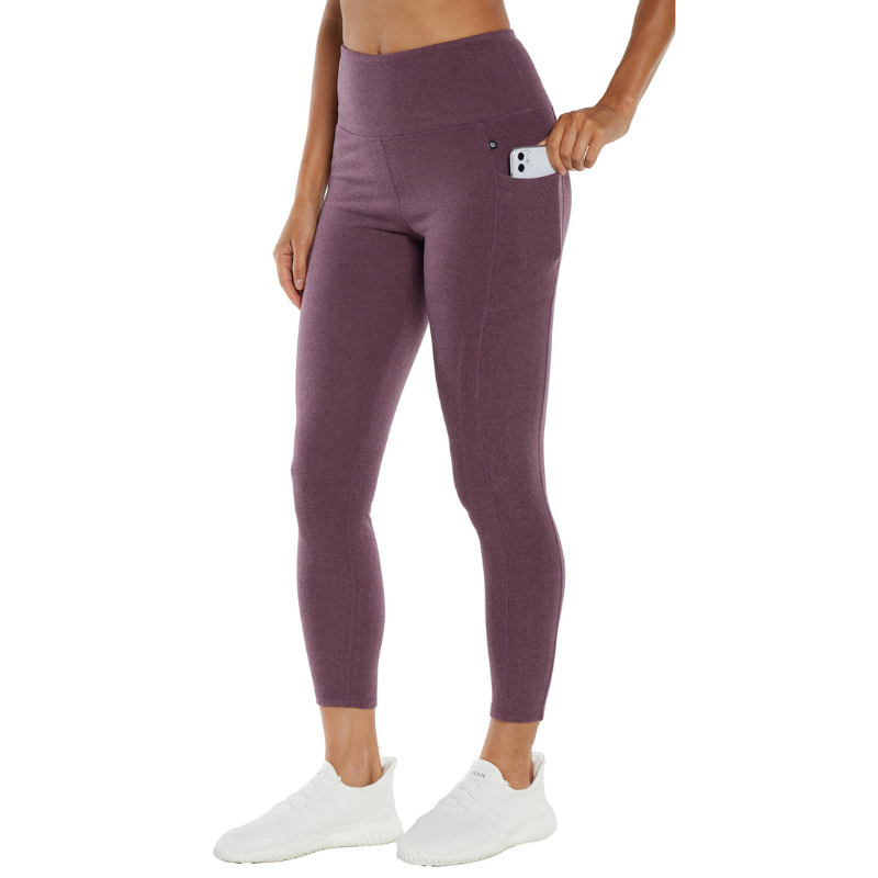 A woman wears high-waisted, purple leggings with a side pocket holding a phone, paired with white sneakers.