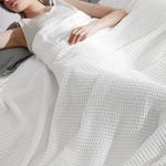 A white waffle weave blanket covers a bed, with a person resting underneath and only their upper body visible.