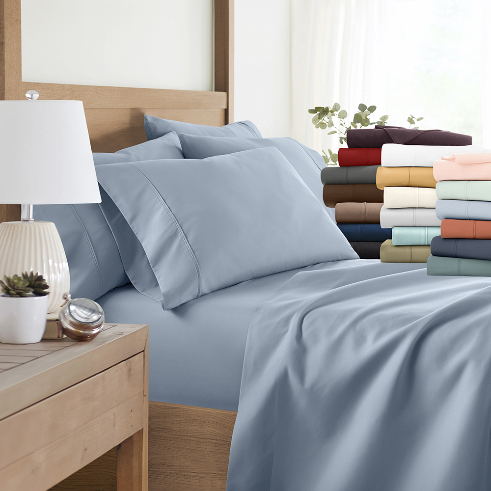 A display of various colored bed sheets and pillowcases, neatly stacked and arranged on a bed with a wood frame and a white lamp on the bedside table.