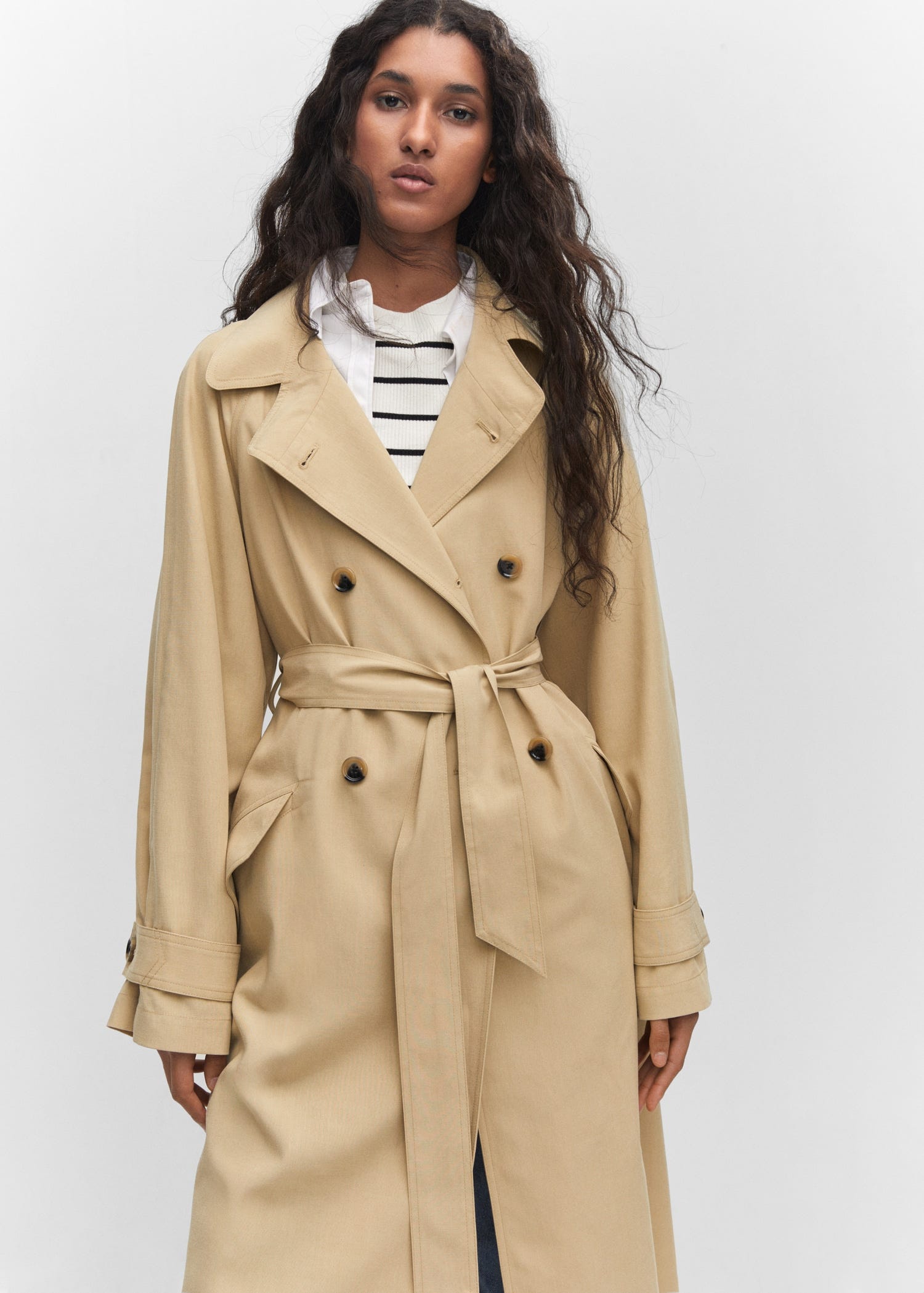 A double-breasted beige trench coat with a belt, featuring epaulettes and buttoned sleeve straps, worn by a model.