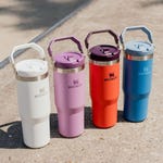 Four insulated drink tumblers in white, purple, red, and blue, each with a handle on the lid, labeled 'Stanley.'