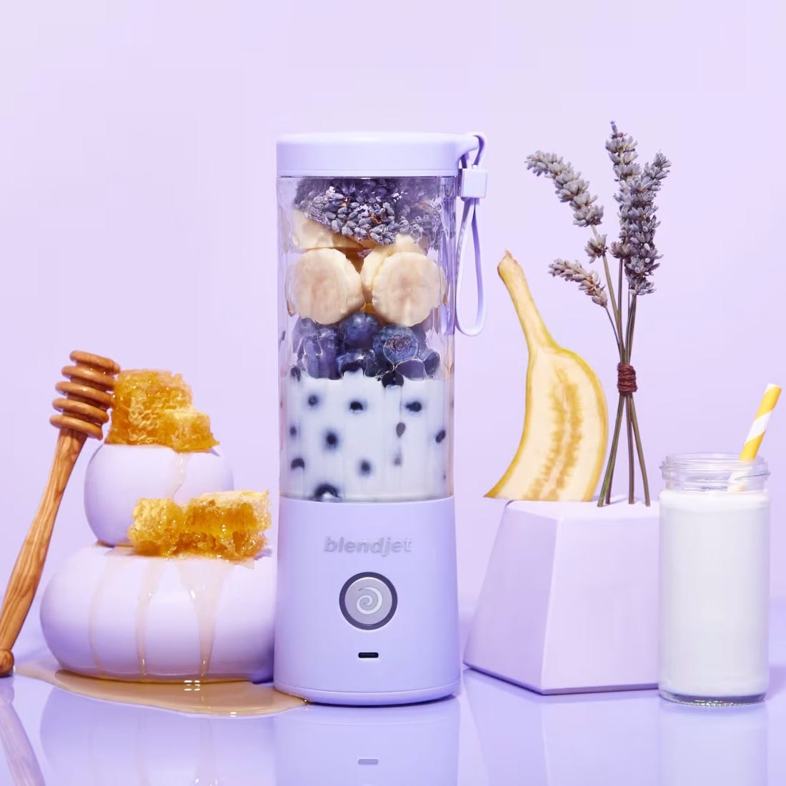 Portable blender with fruits and honey, alongside a banana, milk jar, and lavender sprigs.