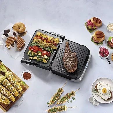 A Cuisinart Griddler is opened to show its dual grilling surfaces, with one side cooking steak and vegetables, and the other toasting sandwiches.