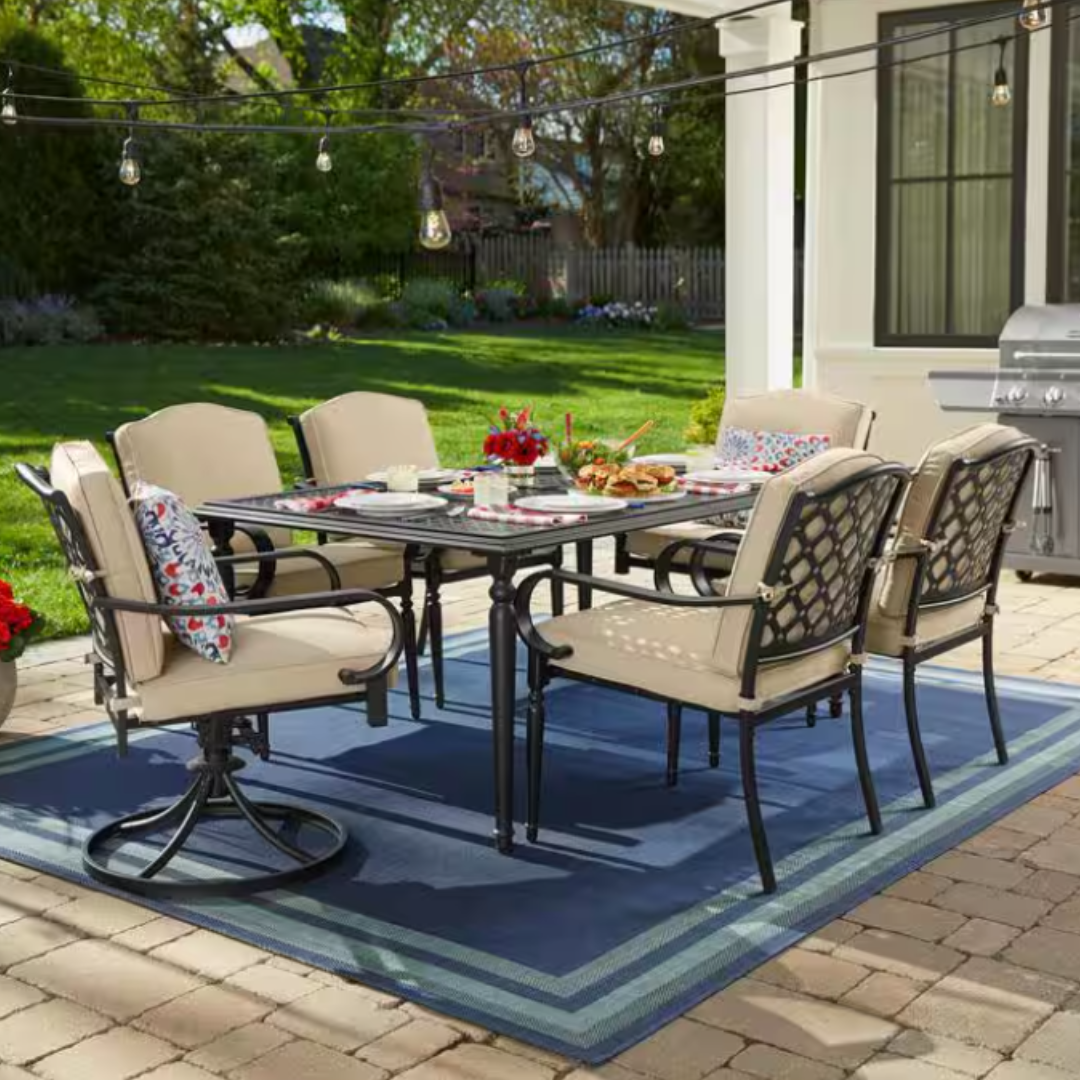 7-piece Hampton Bay Dining Set featuring a rectangular table and six cushioned chairs with beige upholstery. Includes swivel rockers, lattice chair backs, decorative pillows, and is set on a blue outdoor rug; patio string lights and a grill are visible.