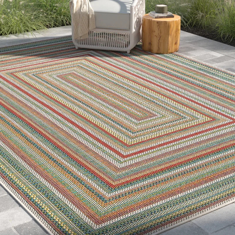 Outdoor patio setup featuring a multi-colored striped rug with a geometric pattern, a wicker chair with a blanket, and a small wooden stump table.
