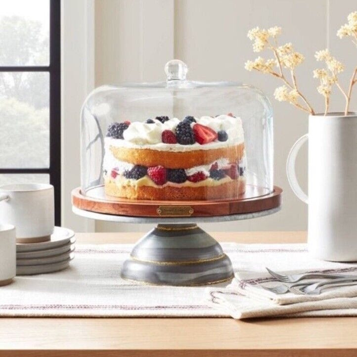 A layered cake topped with berries under a clear glass dome on a wooden stand with a gray base.