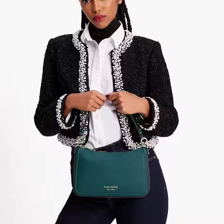 A woman wearing a black textured jacket with pearl trim, a white blouse, and dark trousers, holding a teal shoulder bag.