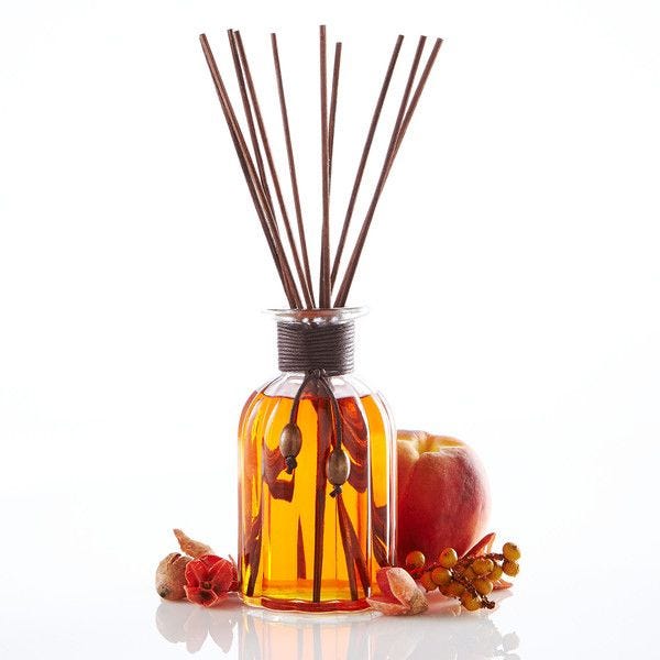 A reed diffuser with several brown sticks in a glass bottle filled with orange liquid, alongside autumnal decorations including an apple and berries.